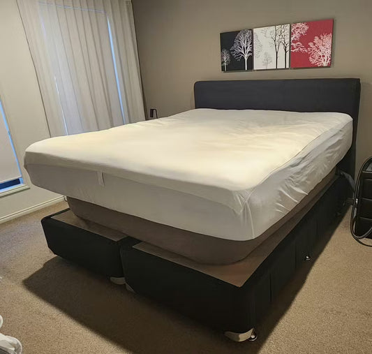 Bedezy installed on an ensemble bed with a connected bedhead. The air hose runs along the head under the mattress and out to the side of the bed where the pump is located behind the lower bed base on it's side.