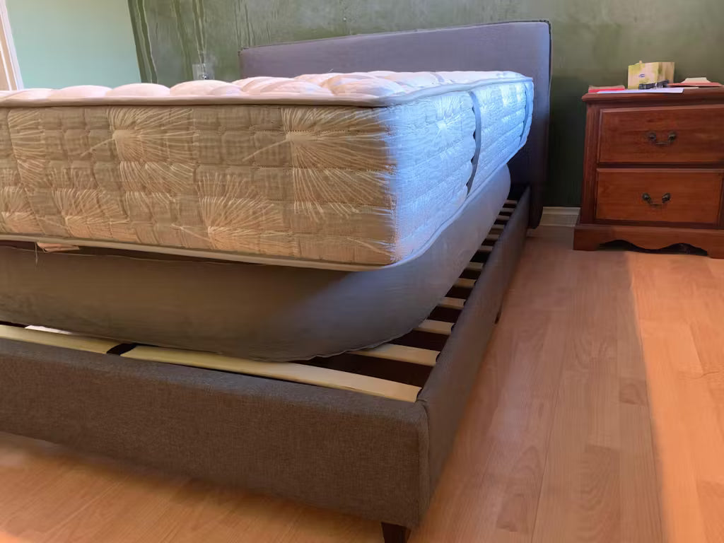 A bed with a mattress on a wooden floor in a room. Bedezy inflated to see the slats on the bed base. The bed base features a side lip where the mattress would be slightly sunken into. 